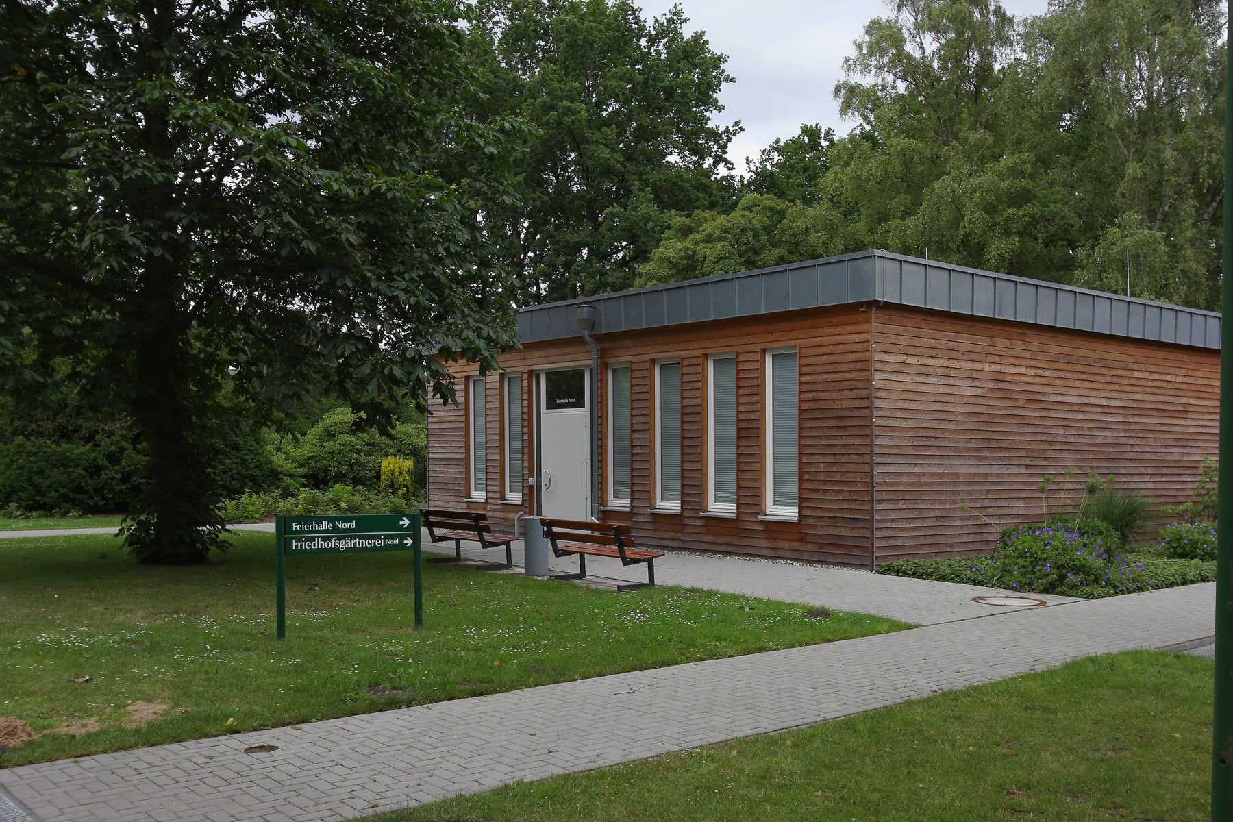 Niedriges Gebäude mit Holzfassade und hohen Fenstern an einem gepflasterten Weg auf Friedhof Öjendorf
